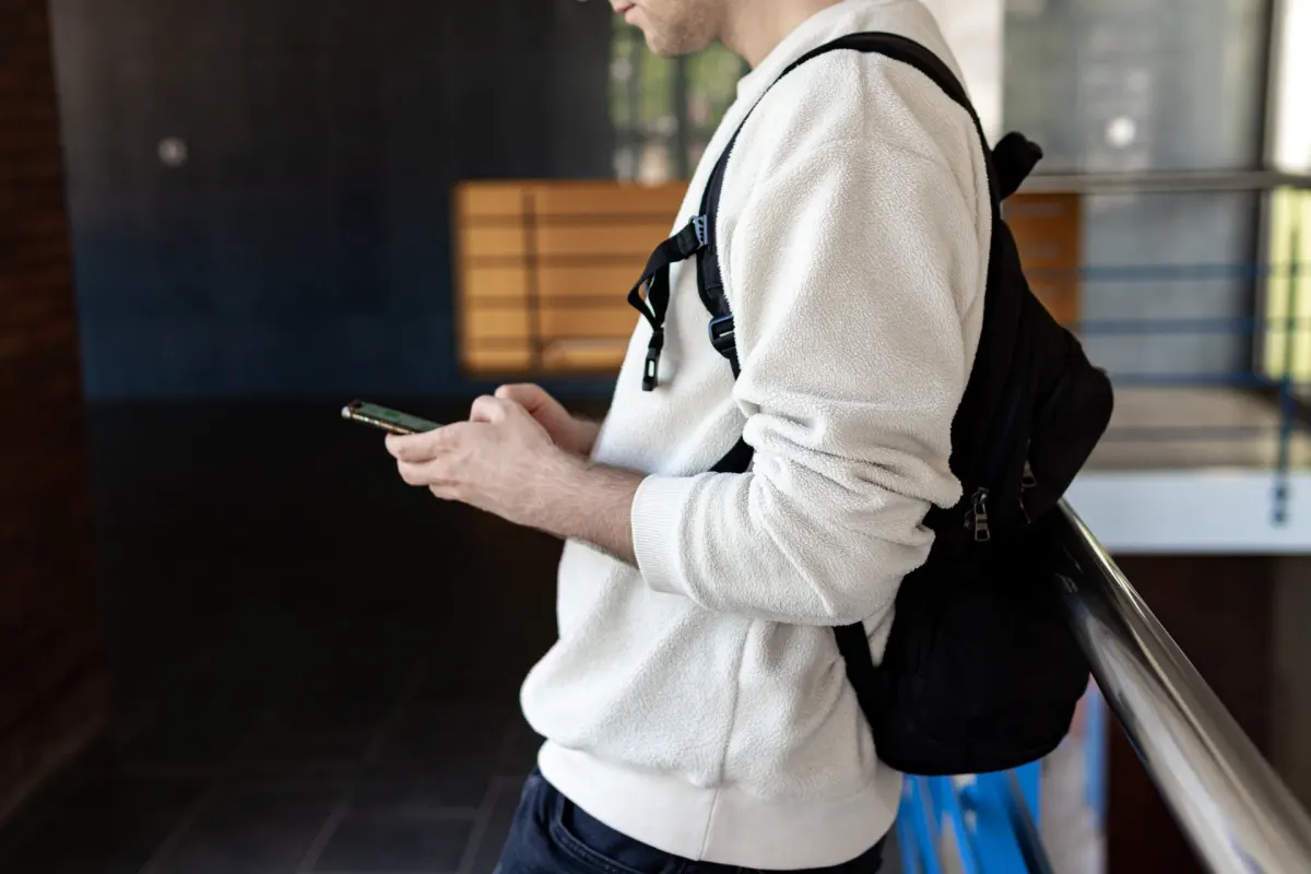 Person in a white jumper and backpack checking a phone by a railing, head out of frame